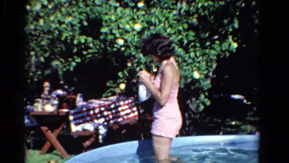 In Ontario, Canada, 1958, the woman looks down at a white bag while standing in a shallow backyard pool near a picnic table.