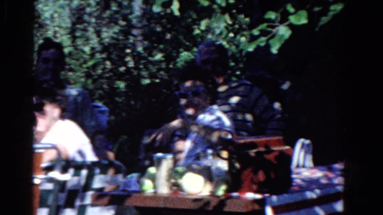 1958 Ontario home-movie frame showing adults gathered at a shaded backyard picnic table with trees and table items around them.
