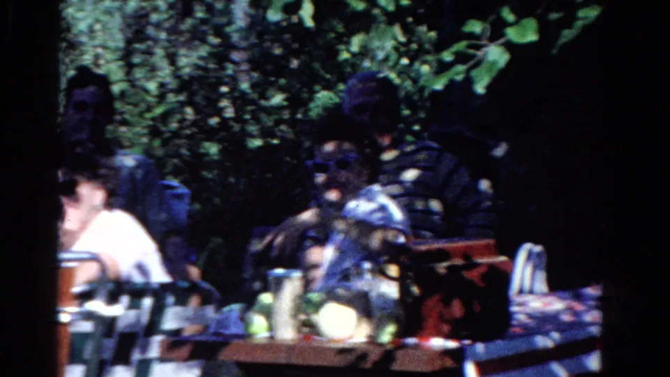 1958 Ontario home-movie frame showing adults gathered at a shaded backyard picnic table with trees and table items around them.