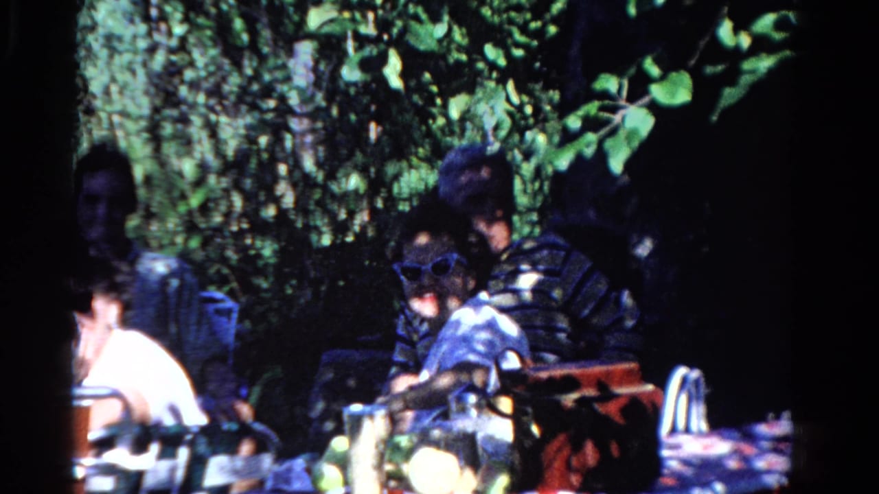 Adults sit at an outdoor table under trees, including a man in sunglasses near picnic items.