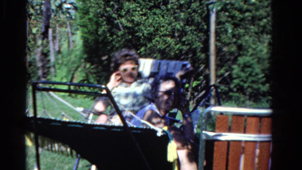 Two adults sit in folding chairs at a wooded campsite.