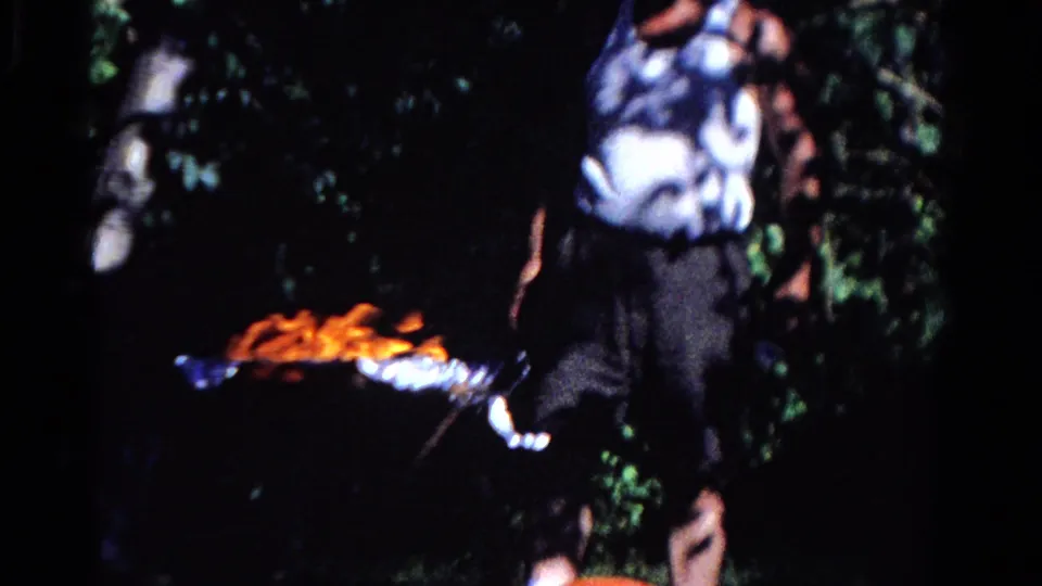 A man in casual sleeveless clothing stands beside a small campsite fire amid dense green foliage.