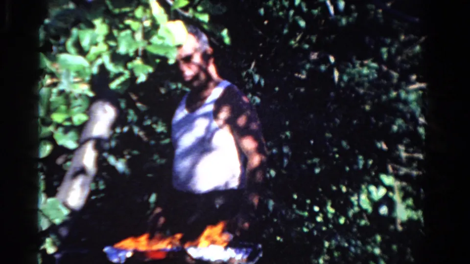 A man wearing sunglasses stands by a small campfire in wooded Ontario home-movie footage from 1958.