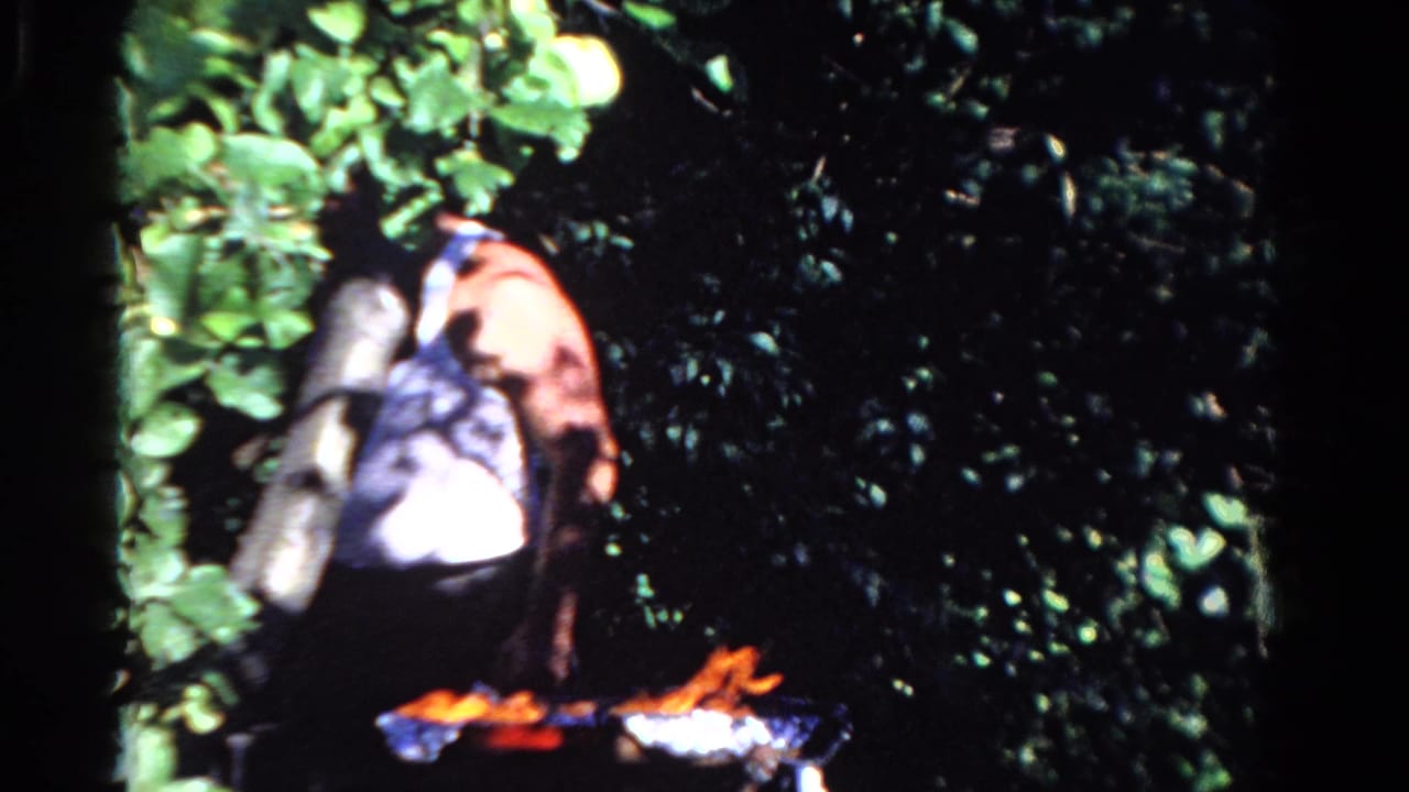 1958 Ontario 8mm frame showing a person beside a small outdoor fire amid leafy greenery.