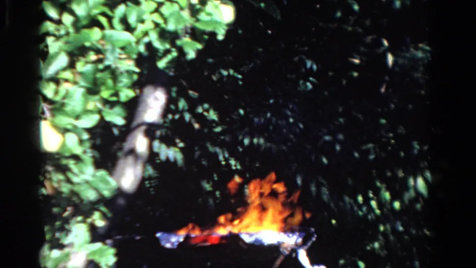 1958 Ontario home-movie frame with an outdoor fire burning against dark green foliage.