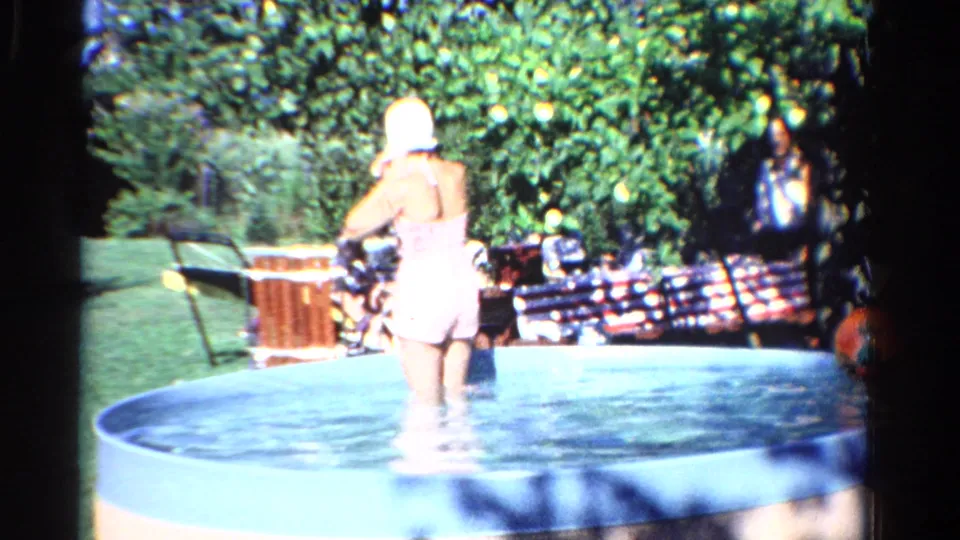 1958 Ontario 8mm frame showing a woman in a pink swimsuit and white swim cap wading in a backyard pool.
