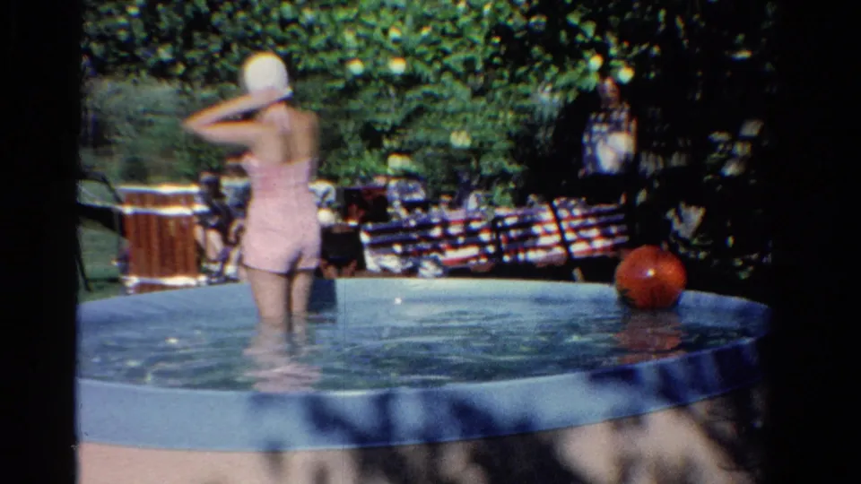 1958 Ontario home-movie frame of a woman adjusting her white swim cap in a pool, with a red ball and picnic setup nearby.