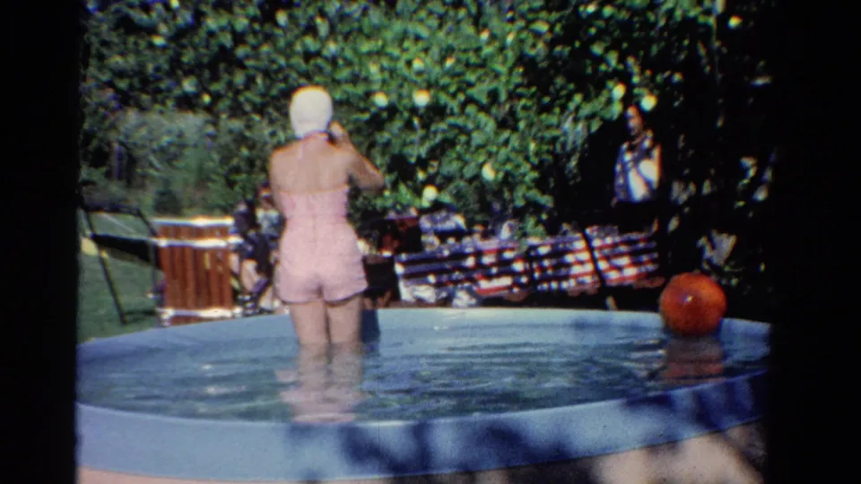 The woman remains in the backyard pool adjusting her white swim cap, with a red ball floating nearby and people seated in shade.