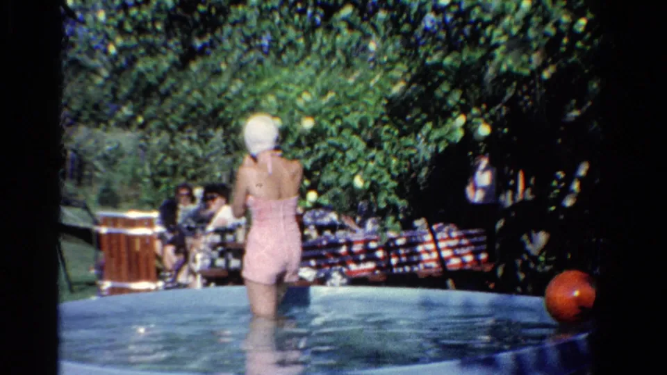 A backyard pool scene shows the woman in a pink swimsuit facing away, with seated people relaxing near a striped blanket beneath trees.