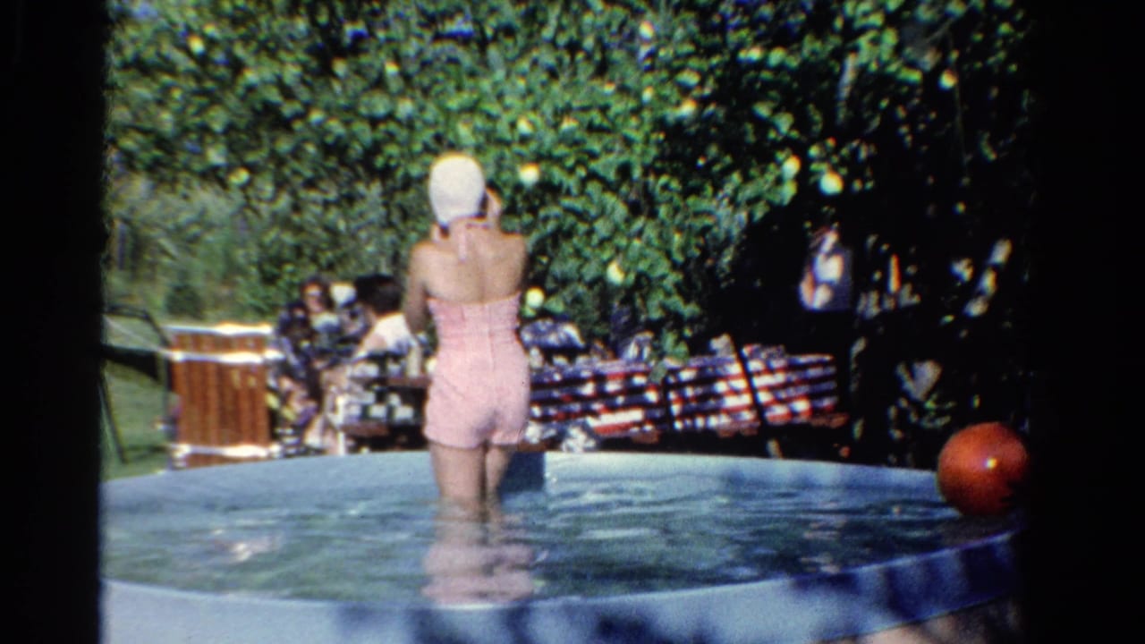 1958 Ontario home-movie frame showing a woman in a backyard pool while people sit under trees nearby.