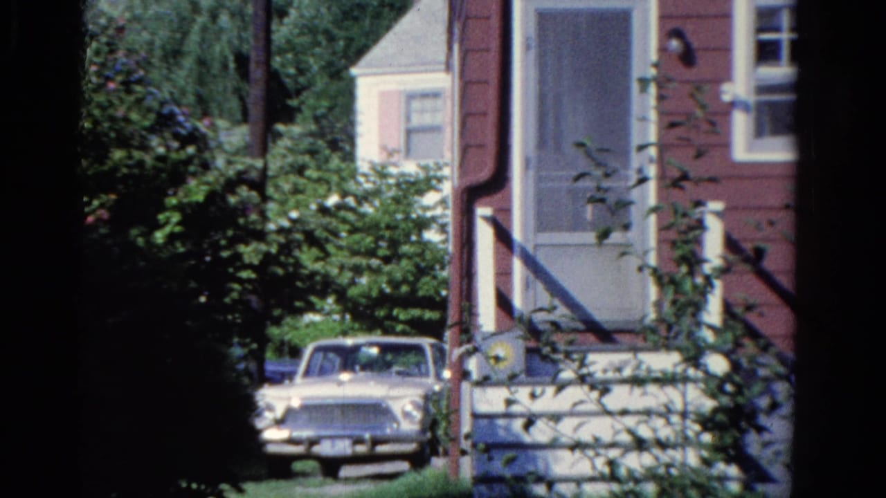 Light-colored 1950s sedan parked beside a red house with white trim and shrubs.
