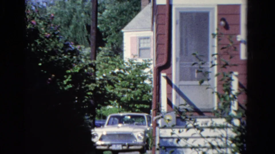 Home-movie view of the Ontario house exterior with a mid-century sedan parked near dense garden shrubs.
