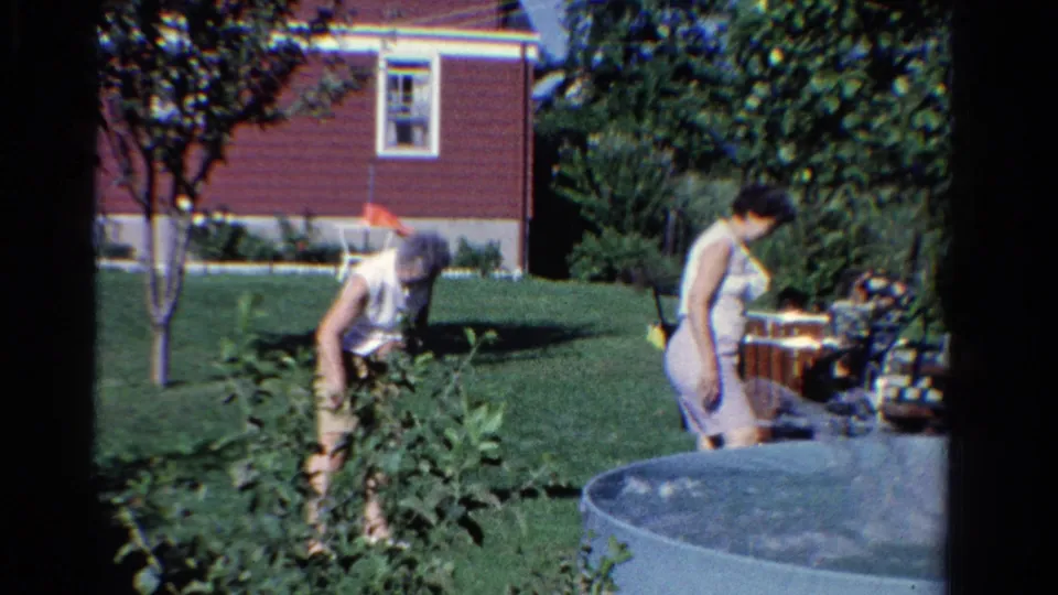 1958 Ontario home-movie frame showing adults near garden plants and an above-ground pool, with a red house and lawn in the background.