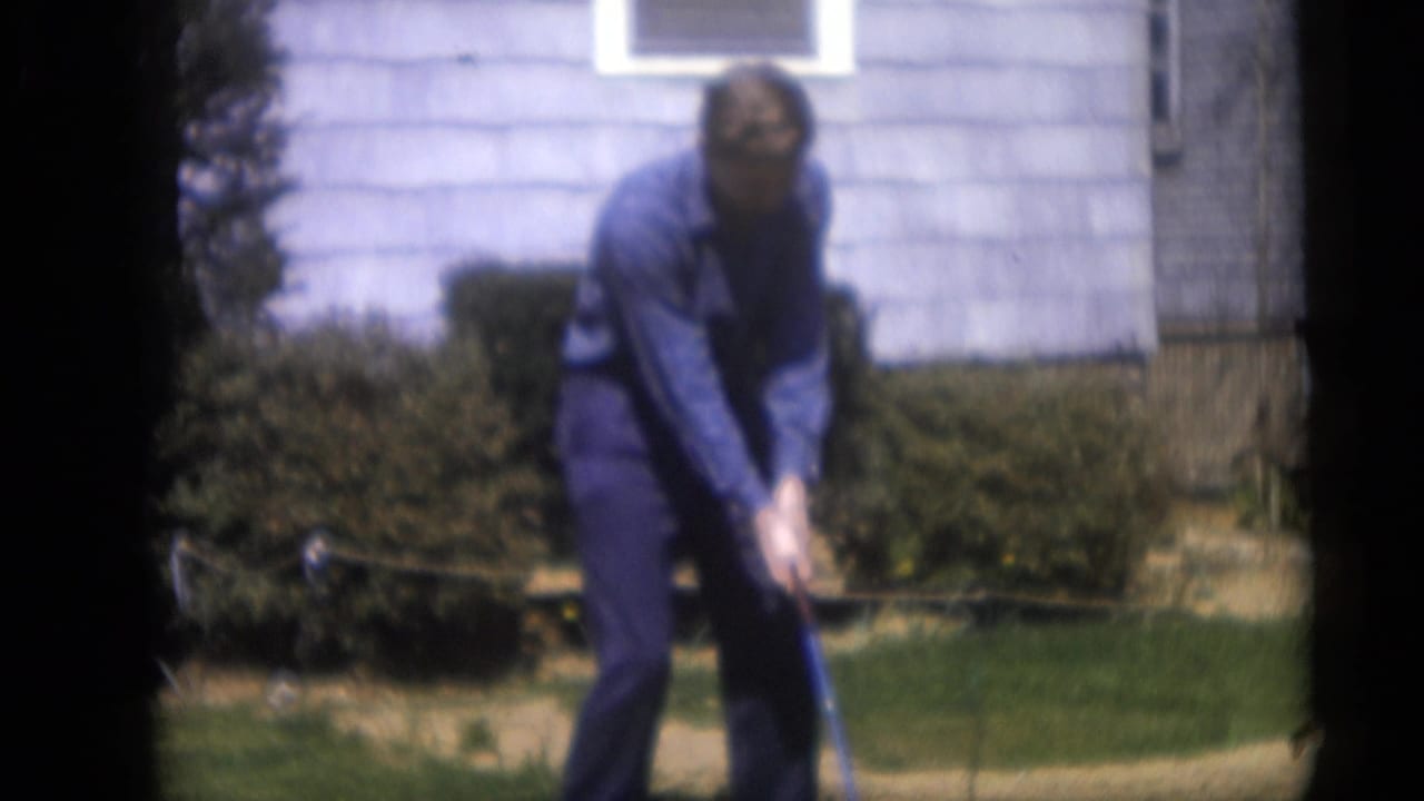 The man remains in his golf stance in the 1959 Camden yard, framed by shrubs, lawn, and a light-colored house exterior.