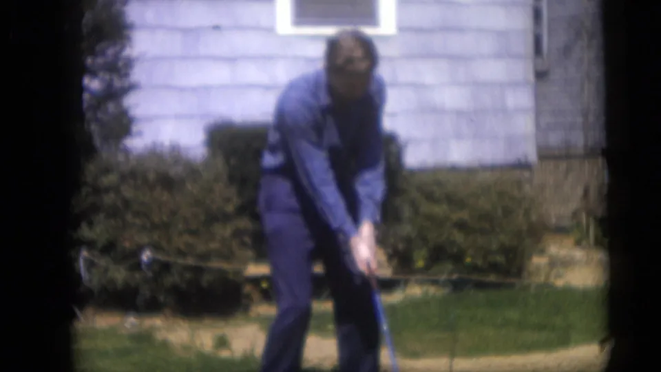 The man remains in his golf stance in the 1959 Camden yard, framed by shrubs, lawn, and a light-colored house exterior.