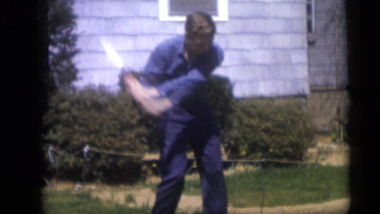 Man bends forward while swinging a golf club in a suburban yard.