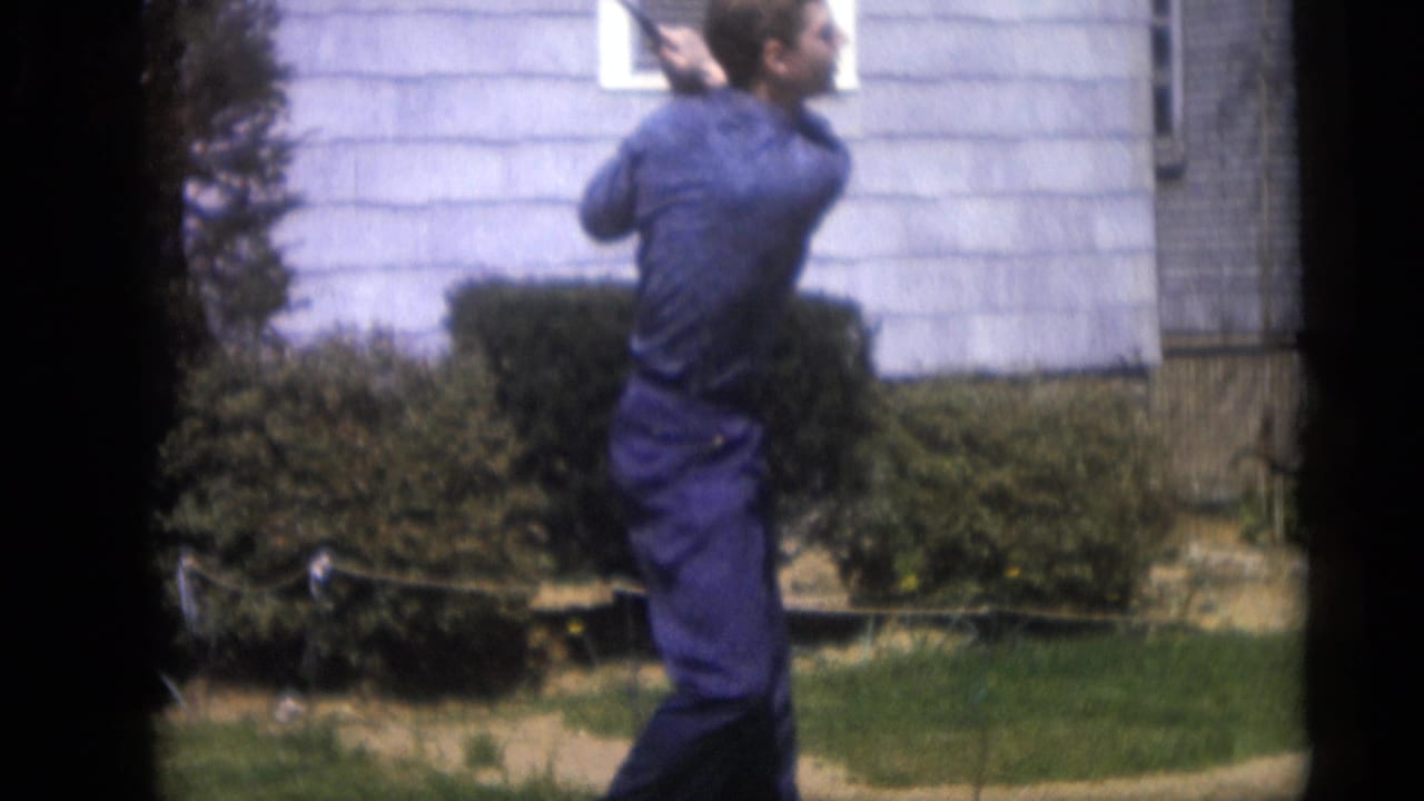 The man finishes the backyard golf swing in front of a light-sided house, captured in soft, grainy 8mm color footage.