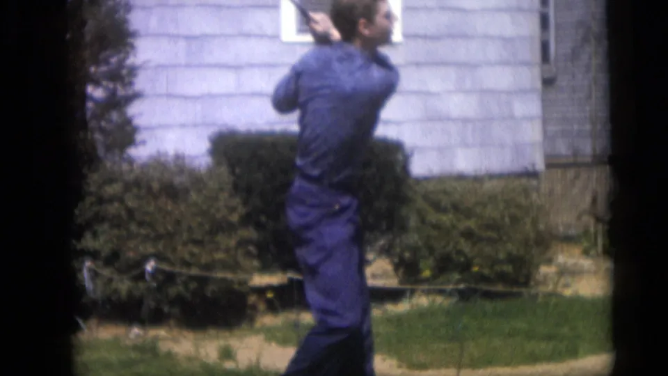 The man finishes the backyard golf swing in front of a light-sided house, captured in soft, grainy 8mm color footage.