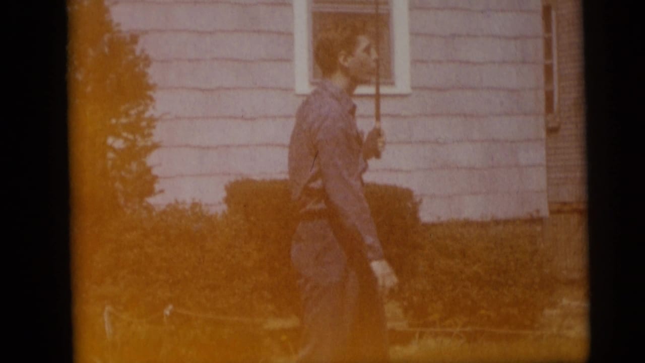 Warm-faded 8mm frame of the man with a golf club in a Camden yard, with house siding, shrubs, and lawn visible.