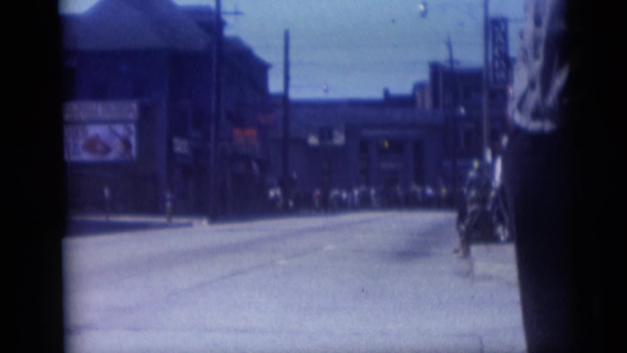 Late frame of the 1959 Camden downtown street view, with commercial buildings, a broad road, utility poles, and distant human activity.
