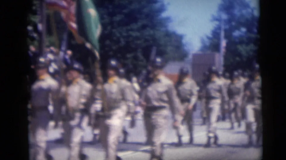 Camden, New Jersey 1959 home movie shows soldiers from behind as flags and a green banner pass through a public parade.