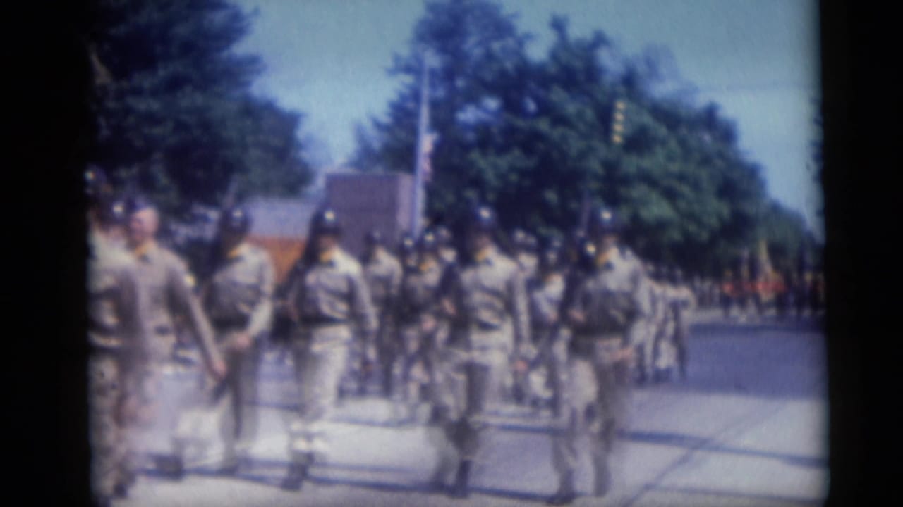 Uniformed marchers in light clothing and dark helmets pass along a sunny street.