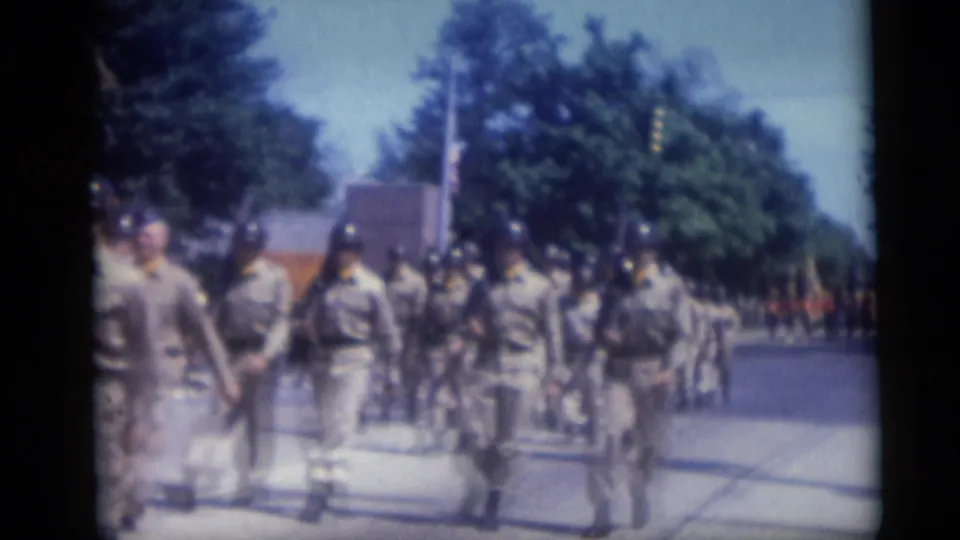 Uniformed marchers in a 1959 Camden parade move down a tree-lined street as spectators watch from the sidewalk.