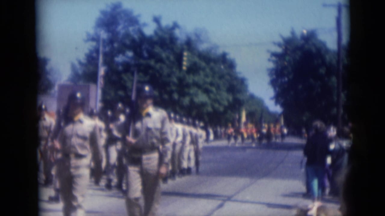 A marching formation continues down the street with spectators along the curb.
