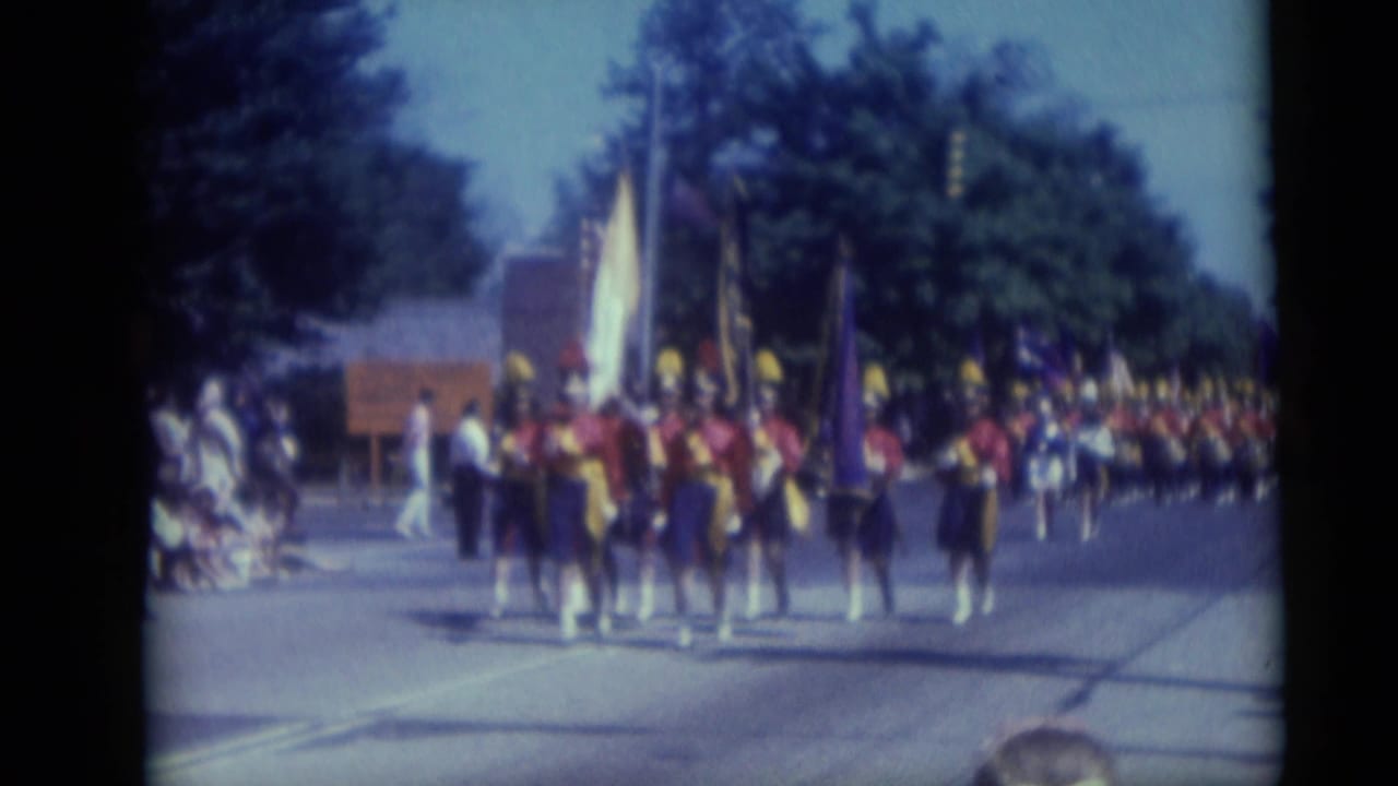 Band members and flag bearers proceed along a Camden street in a late-1950s parade, filmed from a spectator viewpoint.