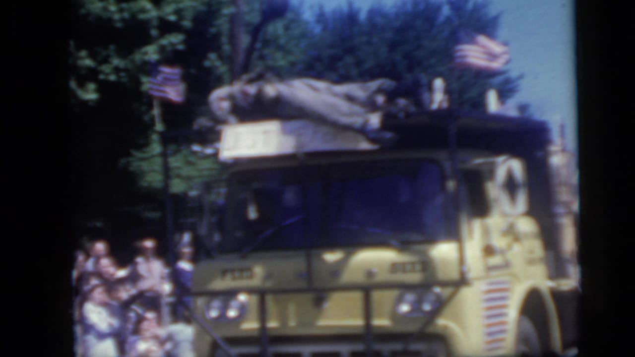 Yellow parade truck with American flags passing spectators under trees.