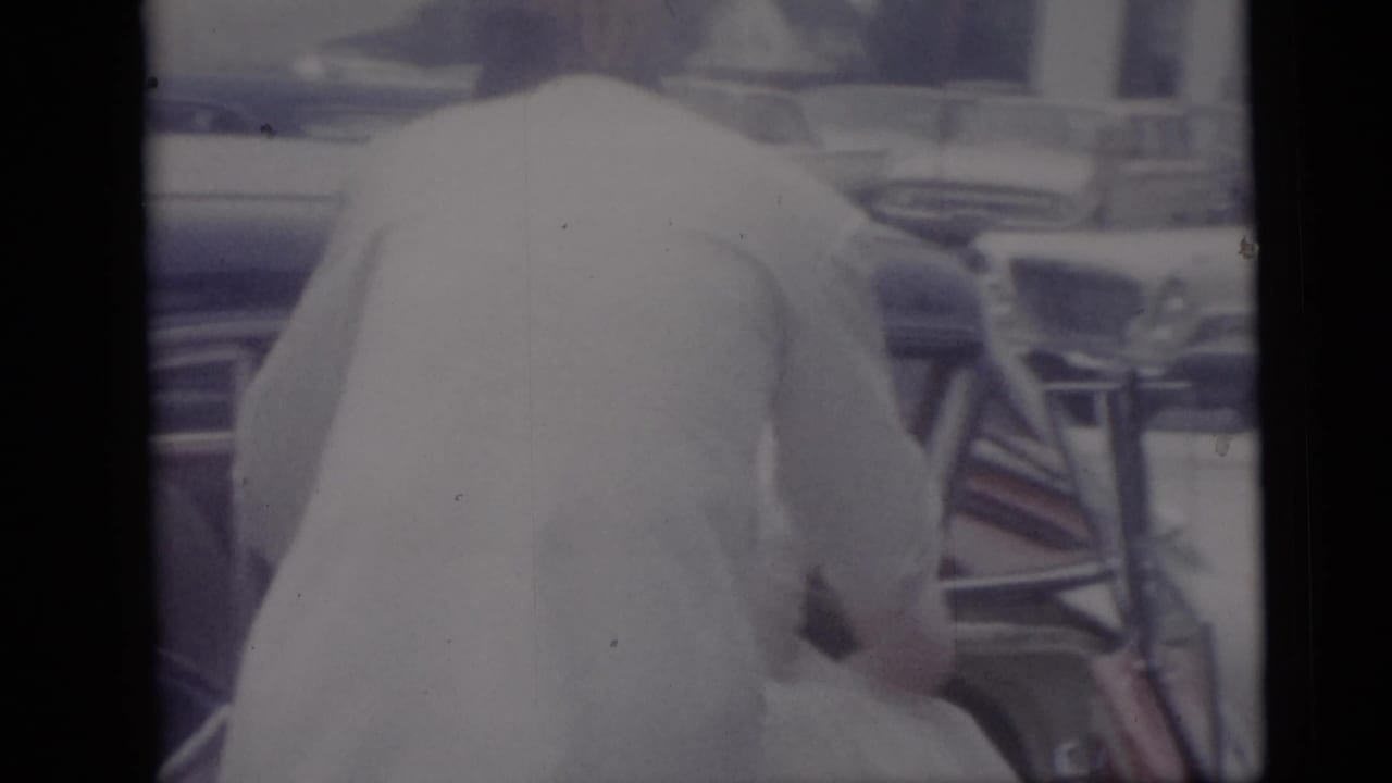 A wedding participant in a light jacket leans near a car, with chrome and interior details visible in a 1959 Camden home-movie frame.