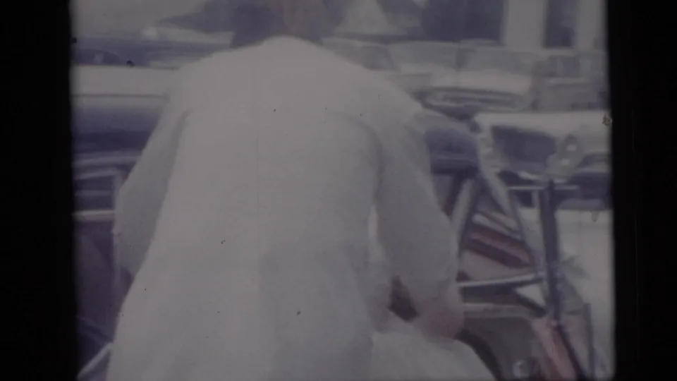 A wedding participant in a light jacket leans near a car, with chrome and interior details visible in a 1959 Camden home-movie frame.