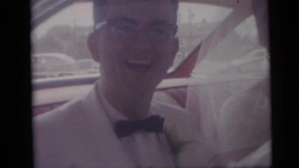 Smiling groom in a light suit and bow tie stands beside a bride in white during a 1959 Camden, New Jersey wedding home movie.