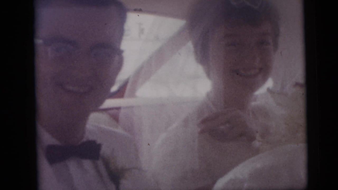 Bride and groom smiling inside a car, seen through a window.