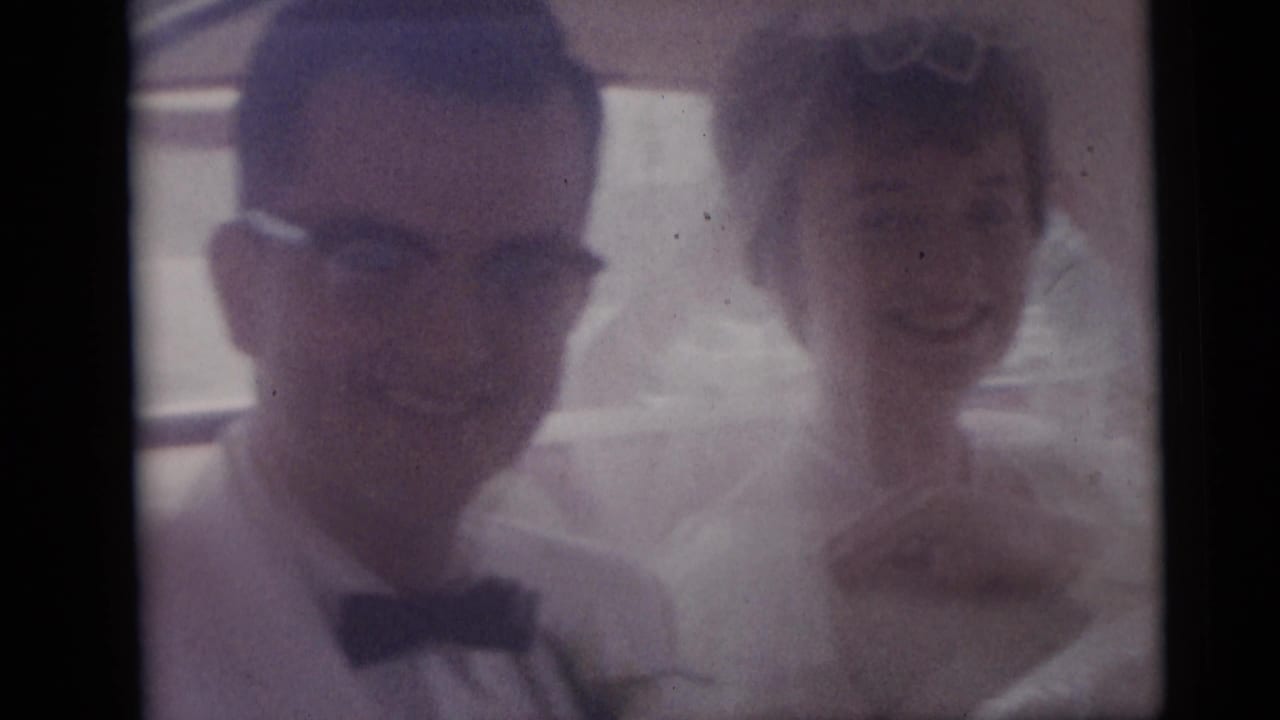 The groom, wearing glasses and a bow tie, sits next to the bride inside the car during the Camden wedding footage.