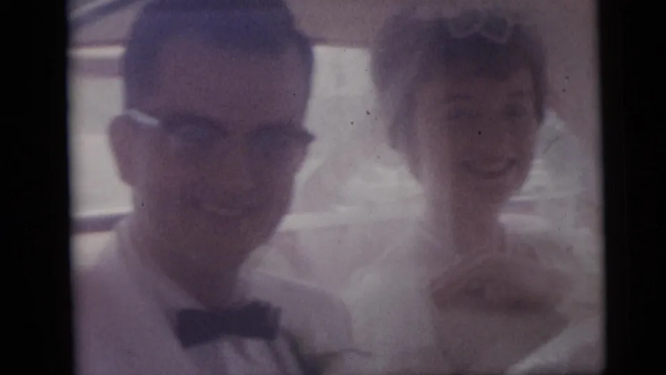 The groom, wearing glasses and a bow tie, sits next to the bride inside the car during the Camden wedding footage.
