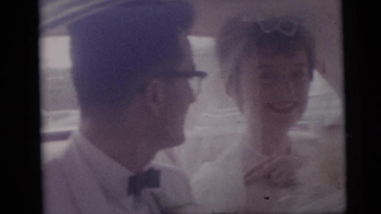 The groom turns toward the bride as she smiles inside the wedding car, with soft window glare across the frame.