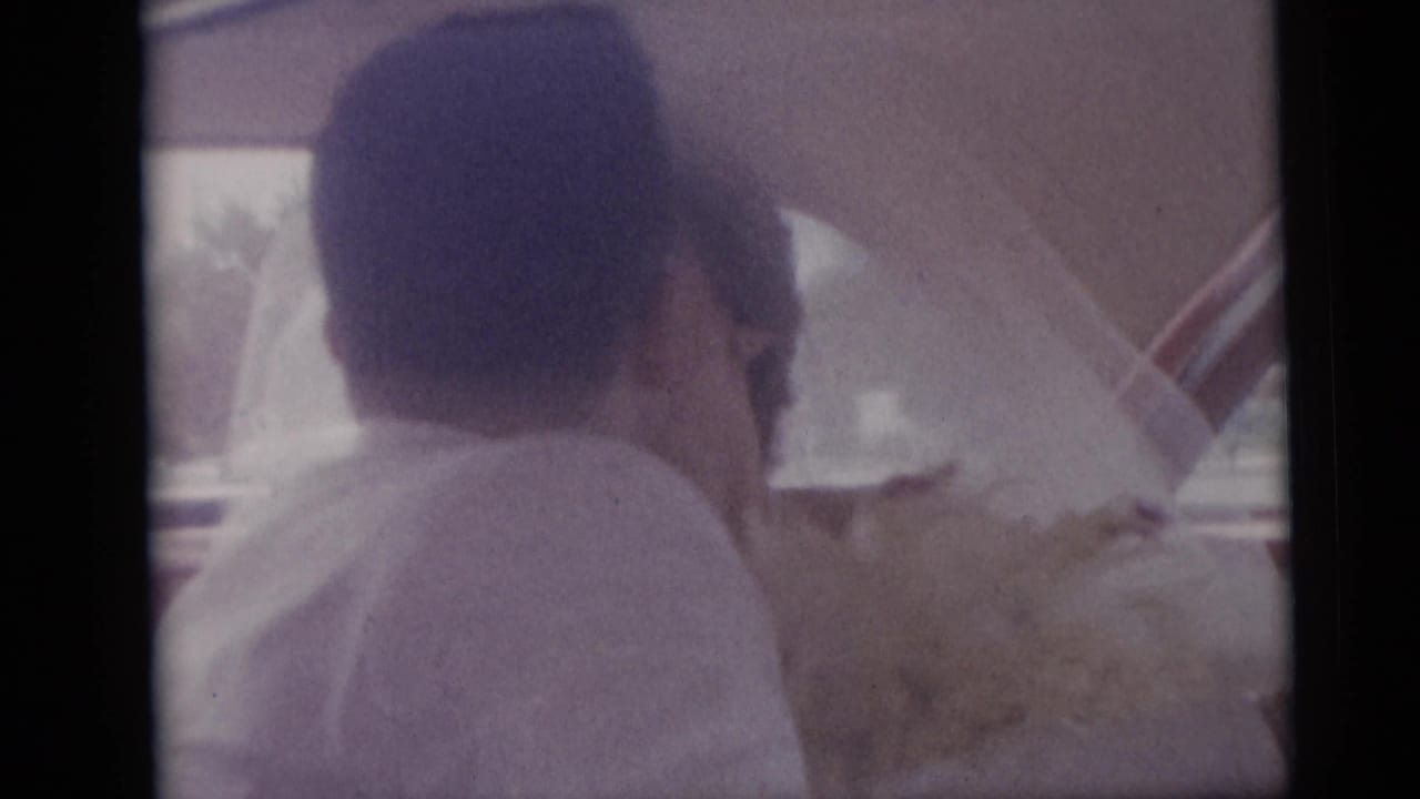 Bride in veil seated in a car, seen through the window beside a bouquet.