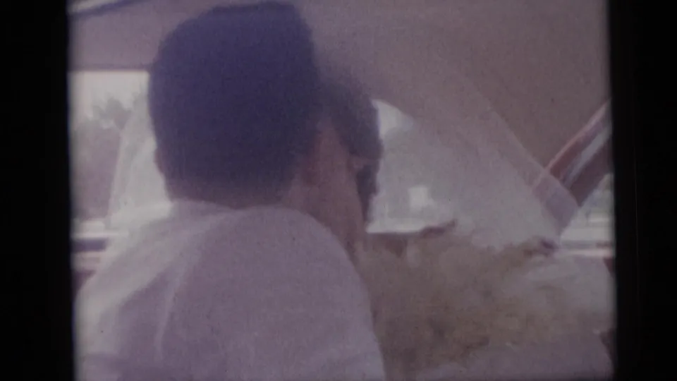 1959 Camden wedding home movie frame showing the bride in a veil seated inside a car with flowers nearby.