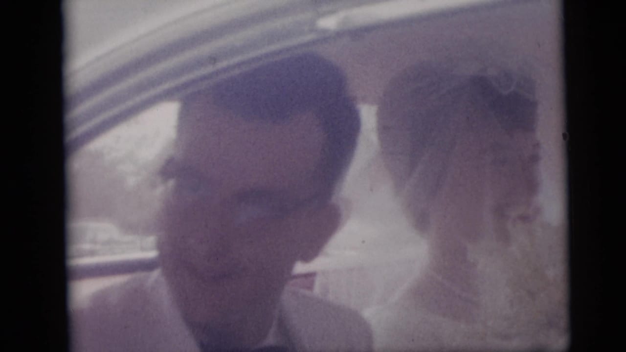 Groom in glasses and bride in veil seated together inside a car.