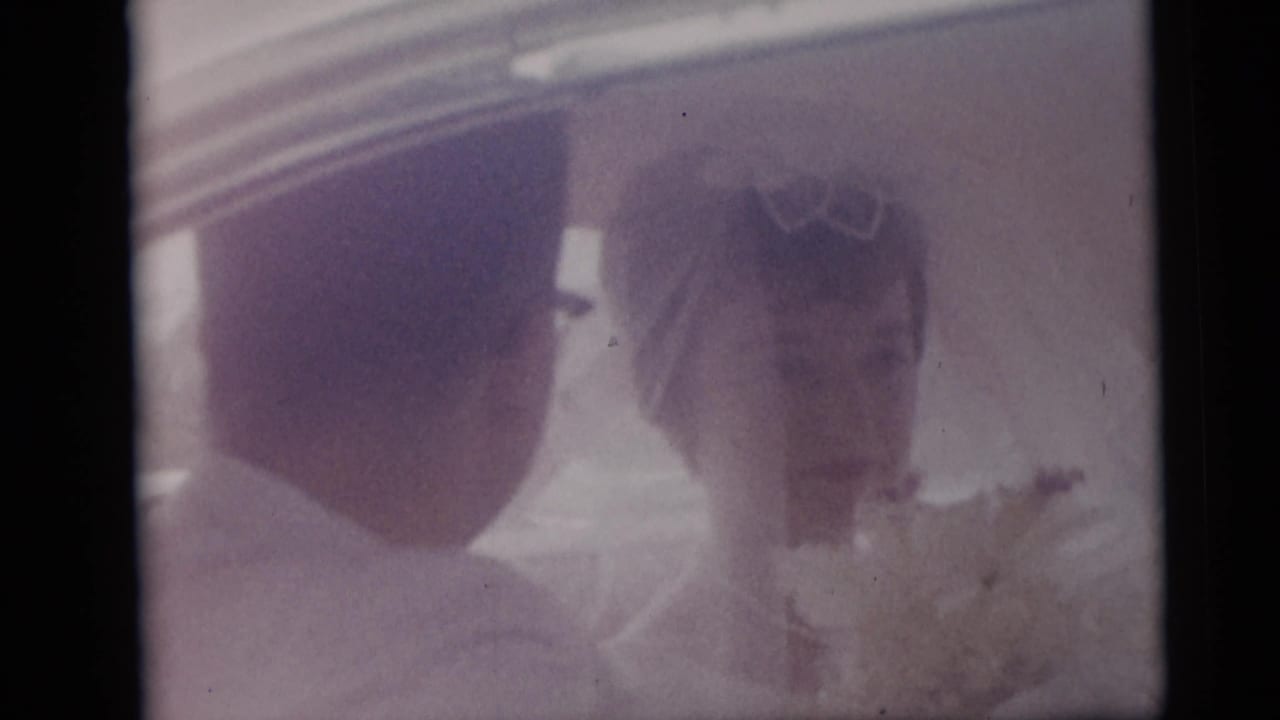 Newlyweds in a 1959 Camden home movie share a close moment inside a car, with the bride's bouquet visible.