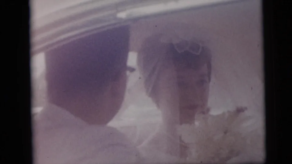 Newlyweds in a 1959 Camden home movie share a close moment inside a car, with the bride's bouquet visible.