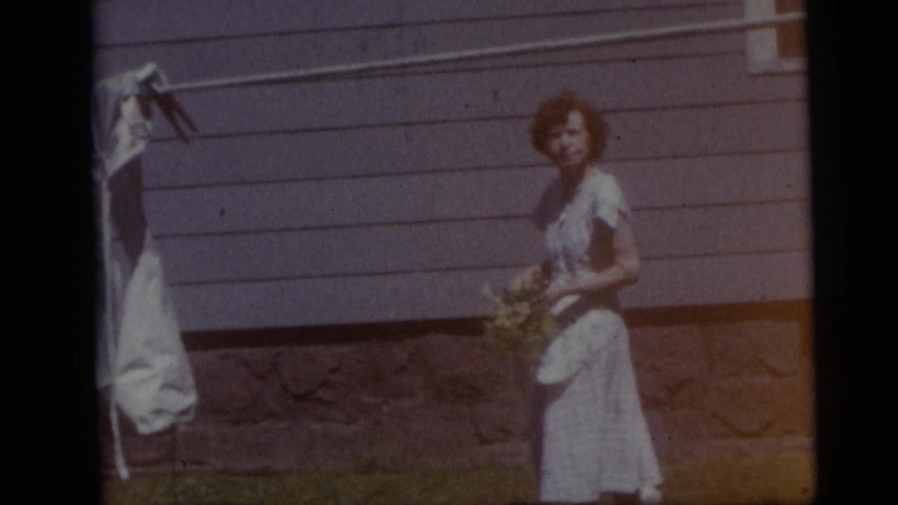 In the 1959 Camden home-movie footage, a woman in a light dress stands near house siding and a clothesline at the edge of a yard.