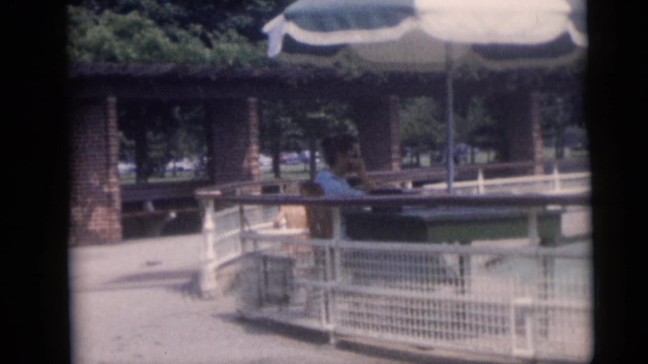 1959 Camden poolside home-movie frame showing white railings, a striped umbrella, brick pavilion columns, trees, and a distant person.
