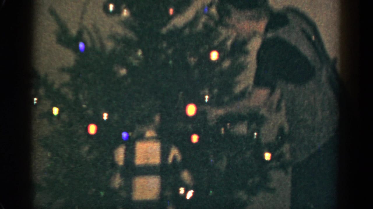 1959 Catskill, New York 8mm frame showing a lit Christmas tree, wrapped gifts below, and a person reaching near the branches.