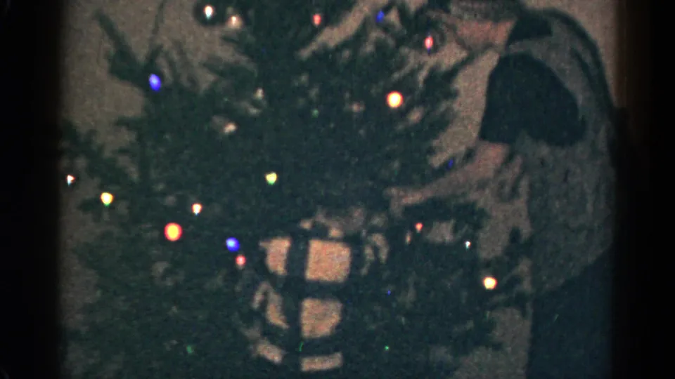 1959 Catskill, New York home-movie frame of a decorated Christmas tree with glowing lights, gifts, and a partially visible adult figure.