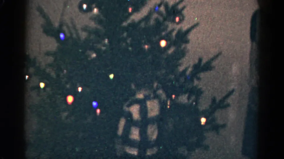 1959 Catskill, New York 8mm frame focusing on a Christmas tree with colored bulbs, ornaments, and a wrapped gift at the base.