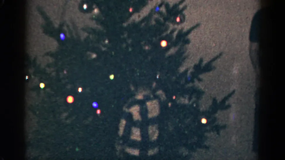 1959 Catskill, New York home-movie frame showing glowing colored tree lights, dense branches, and a wrapped present below.