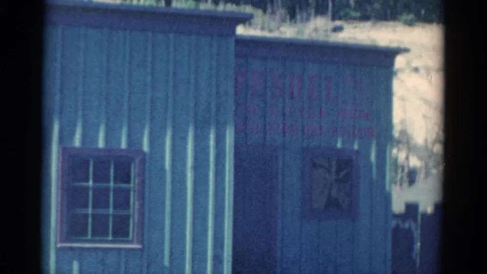 1959 Catskill-area 8mm frame showing blue siding, a barred window, faded red wall text, and a small boarded opening.