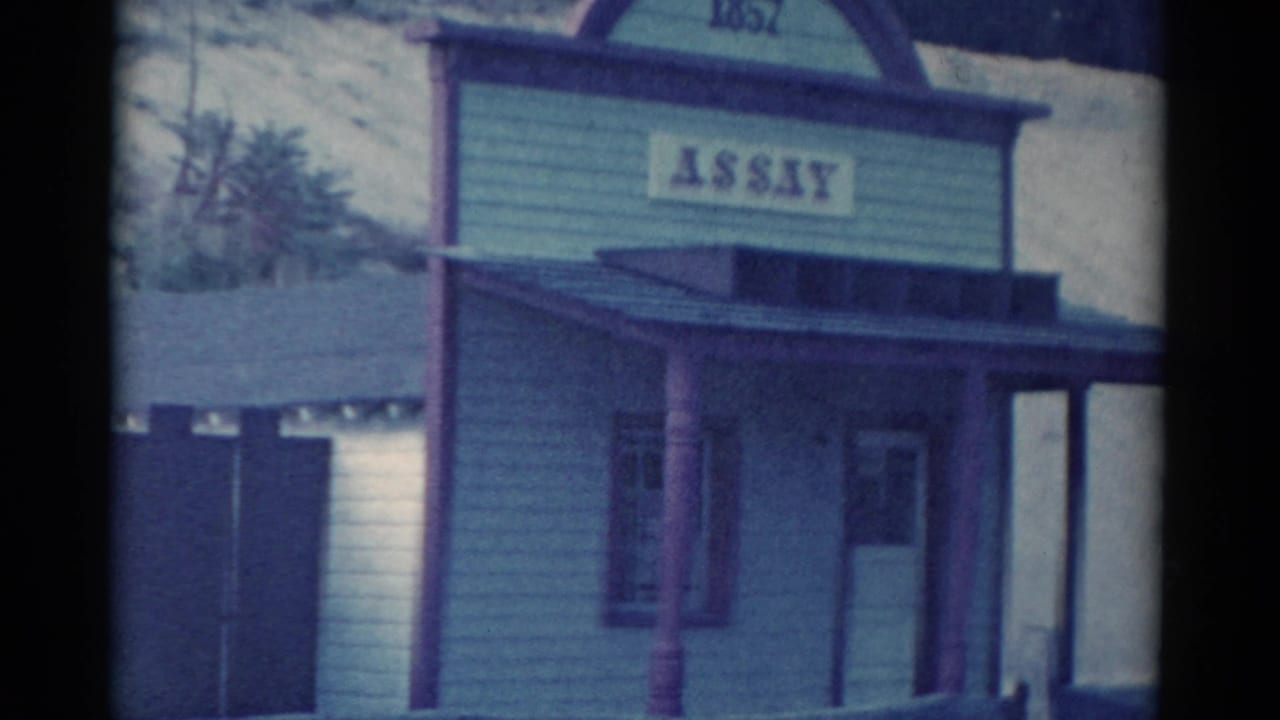 Wooden resort-style building with an ASSAY sign, porch posts, and hillside behind it.